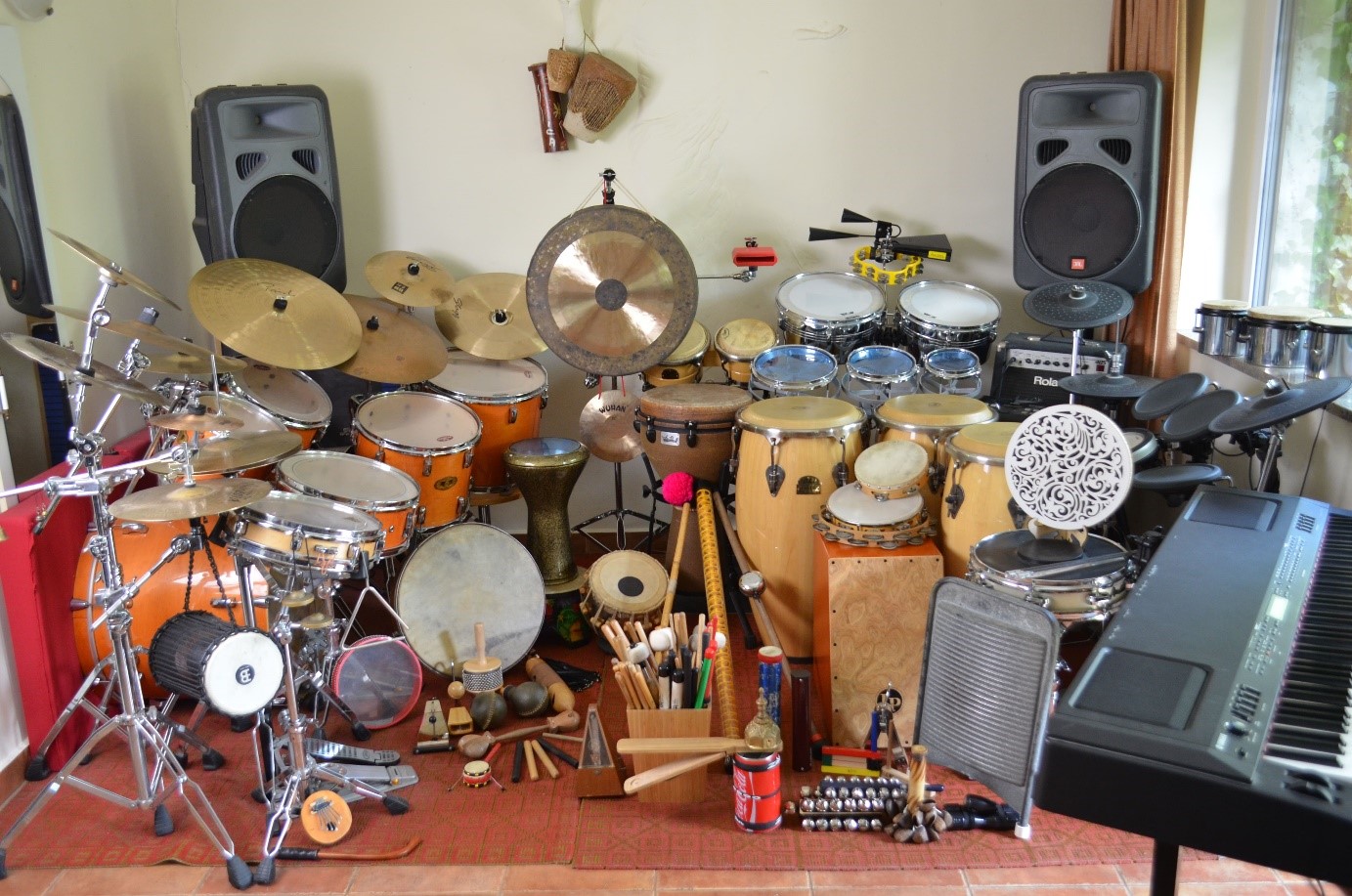 Obraz przedstawiający rozłożone instrumenty na całej szerokości pokoju: zestawy perkusyjne, kongi, talerze, rototomy, cajon, piano i wiele innych.
